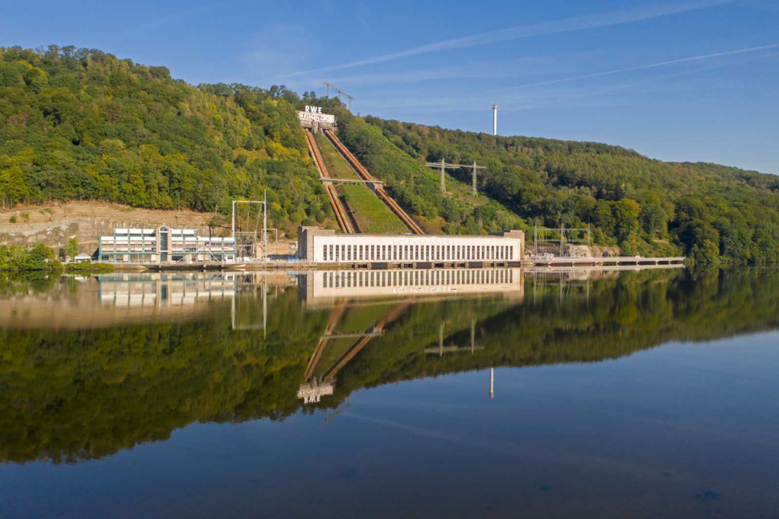 A hydroelectric power station by a calm lake, surrounded by wooded hills, reflecting in the water.