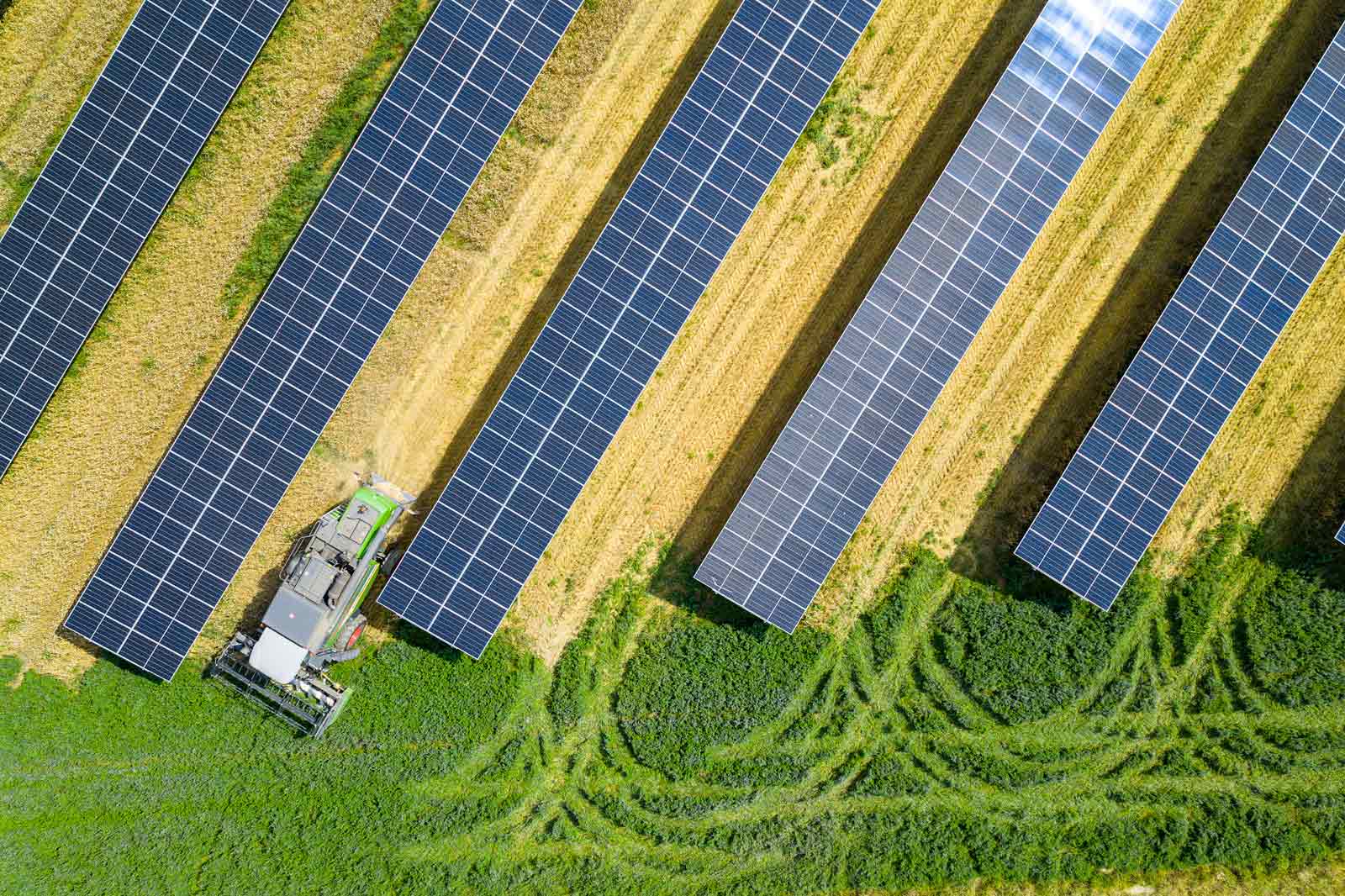 An aerial view of solar panels arranged in rows, with a nearby tractor working in a farmland.