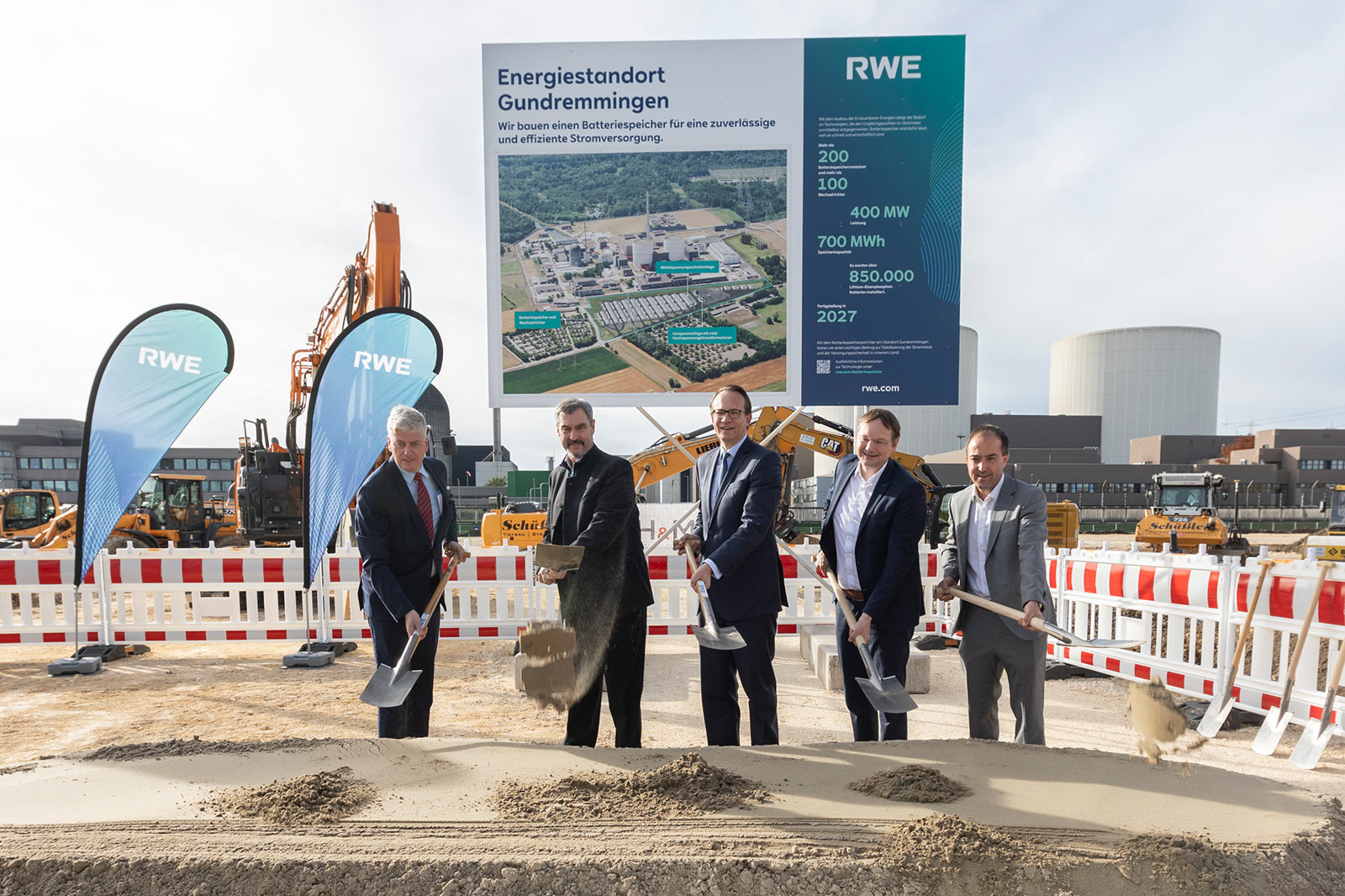 Fünf Personen in Anzügen führen einen symbolischen Spatenstich vor einer Baustelle mit RWE-Fahnen, Baggern, Absperrungen und einem Kühlturm durch.