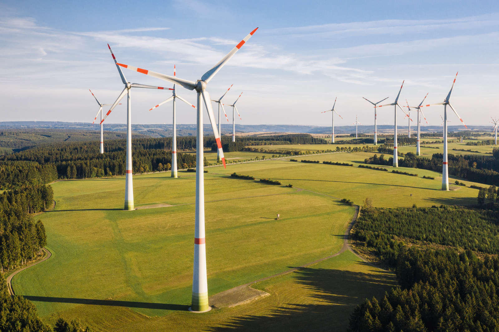 A wind farm with several turbines set in a green landscape under a clear sky, surrounded by forests and fields.