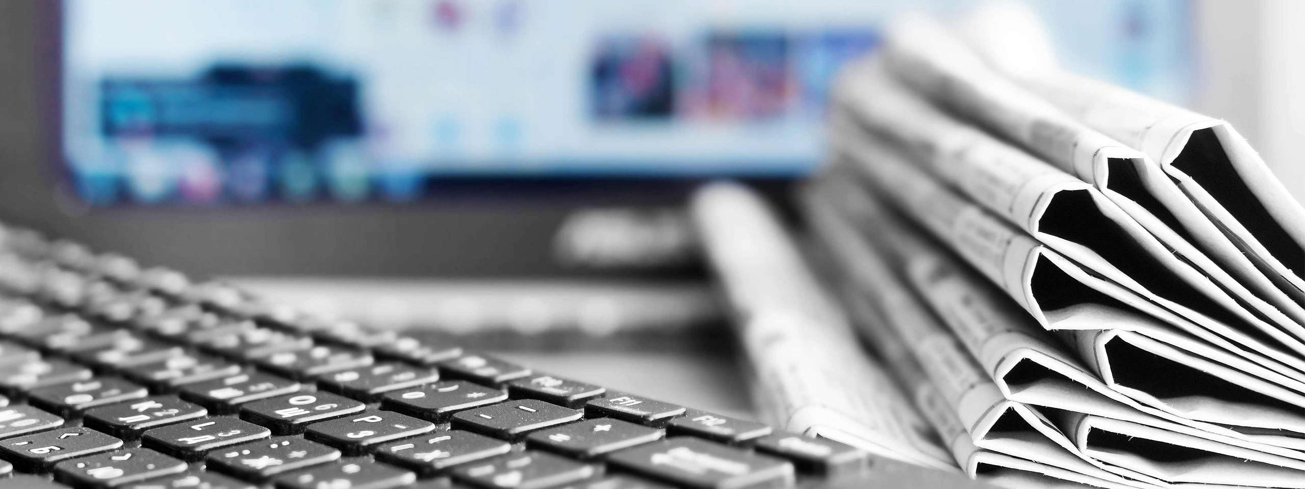 A close-up of a keyboard with newspapers stacked nearby and a blurred computer screen in the background.