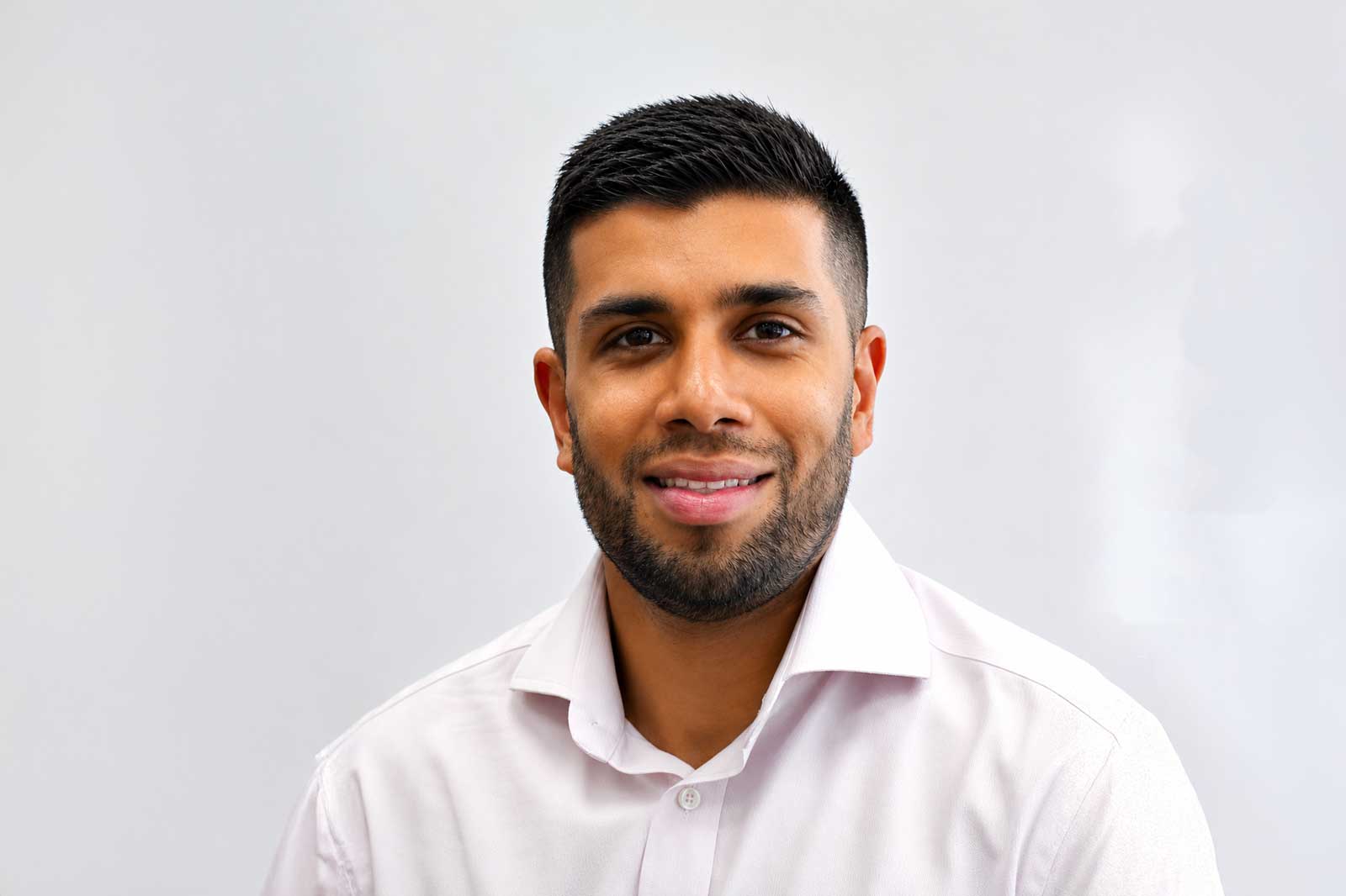 A young man with short dark hair and a trimmed beard smiles warmly while wearing a white collared shirt against a pale gray background.Portrait of Akhil Shah.