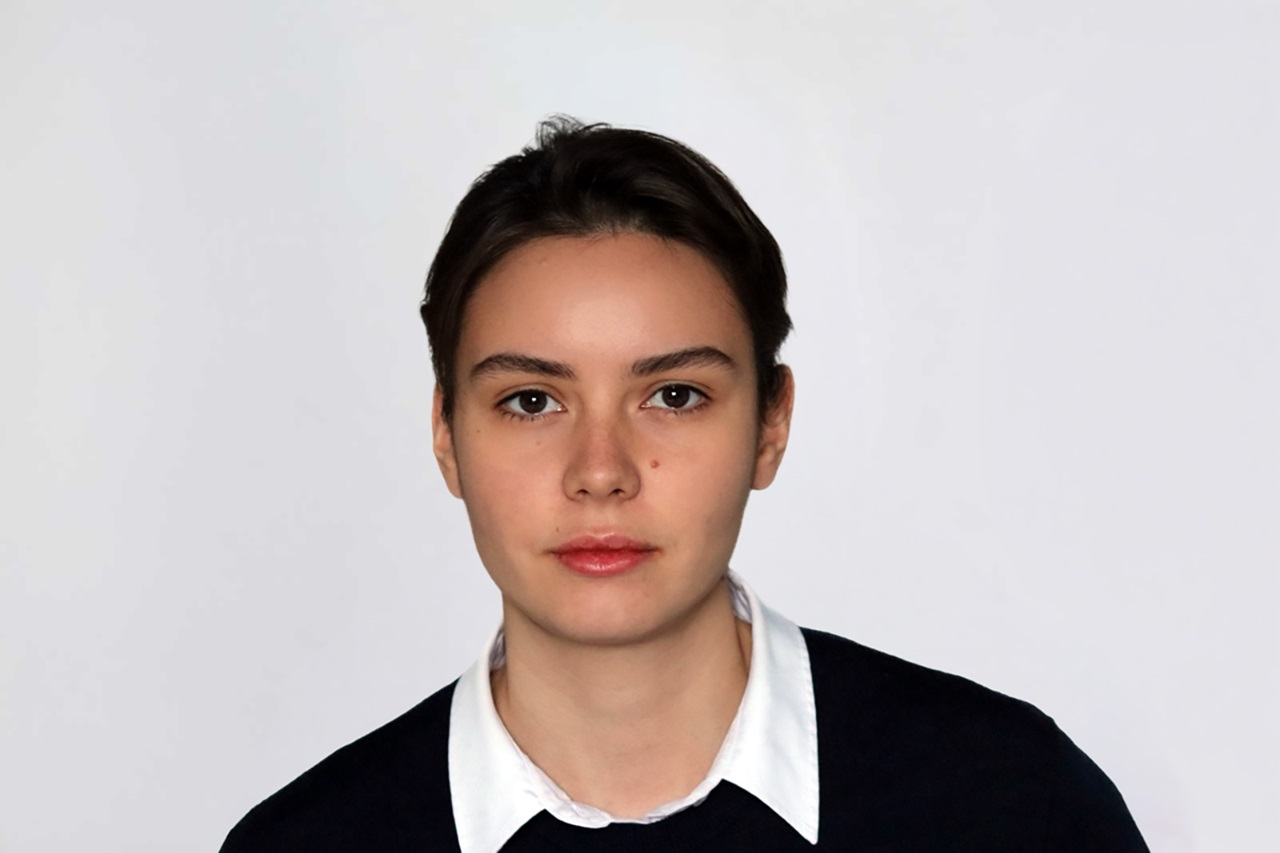 A person with short dark hair and a neutral expression wearing a white collared shirt under a dark sweater against a plain light gray background. Portrait of Bianca Schley.