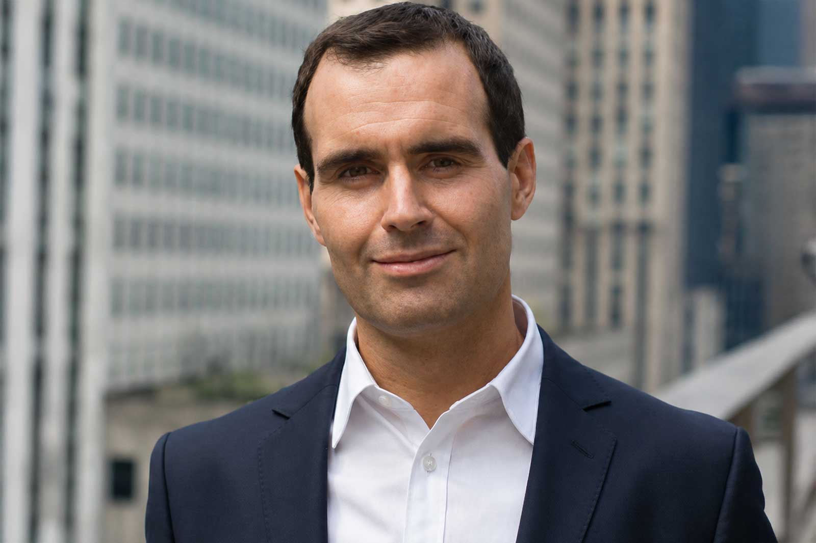 A man in a dark navy suit and white shirt stands on a rooftop terrace with gray skyscrapers behind him, wearing a slight smile. Portrait of Stephen O'Reilly.