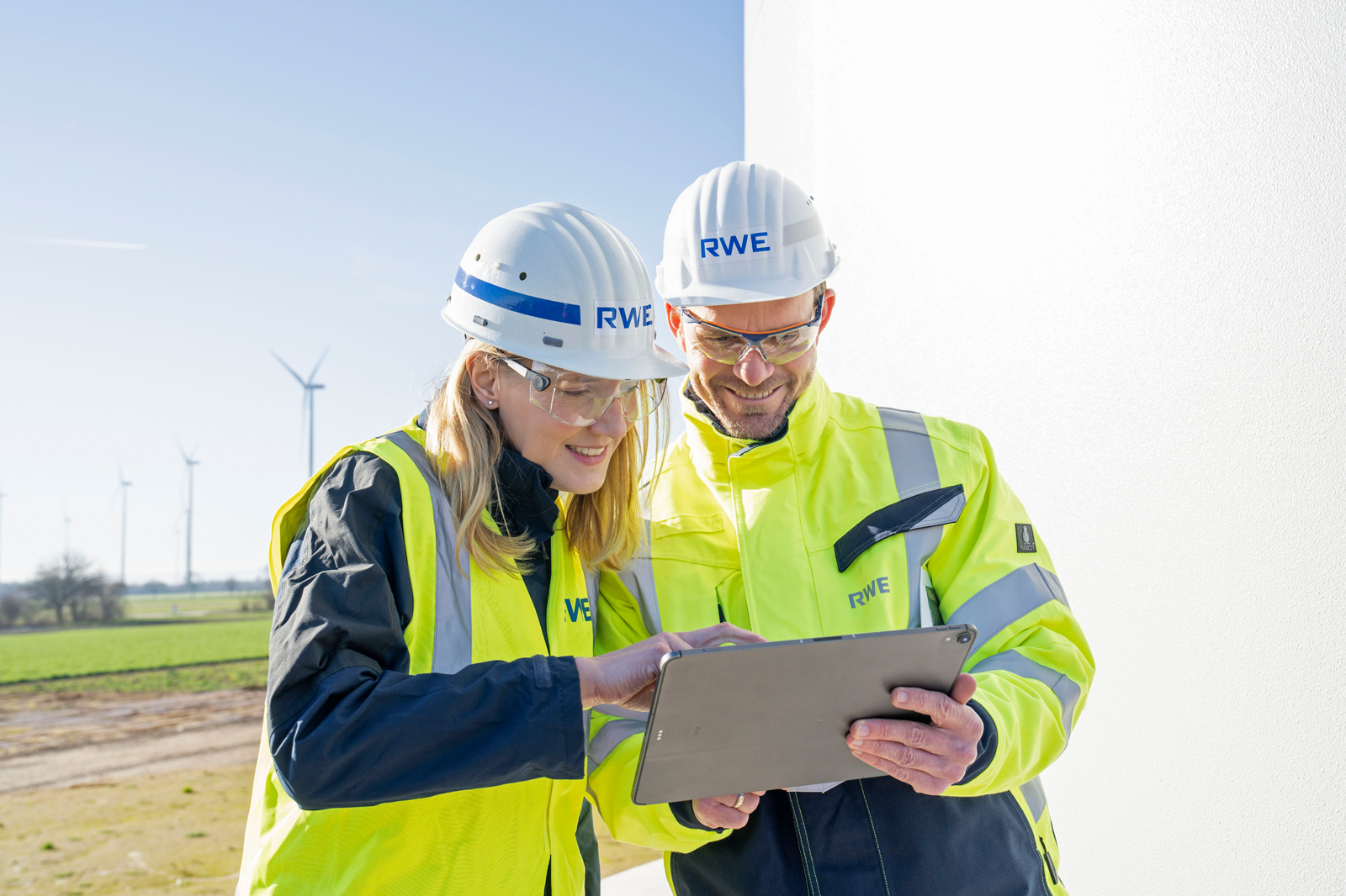 Zwei Arbeiter in Sicherheitskleidung besprechen sich über ein Tablet, mit Windrädern im Hintergrund und klarem blauen Himmel.