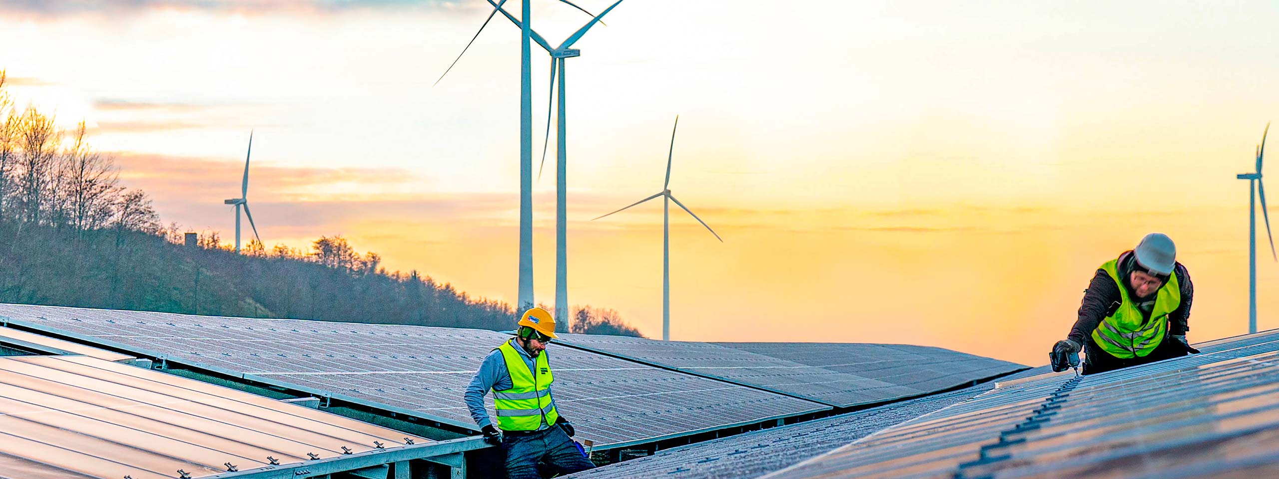 Zwei Arbeiter in Sicherheitskleidung installieren Solarpanels auf einem Dach bei Sonnenuntergang, mit Windturbinen im Hintergrund.