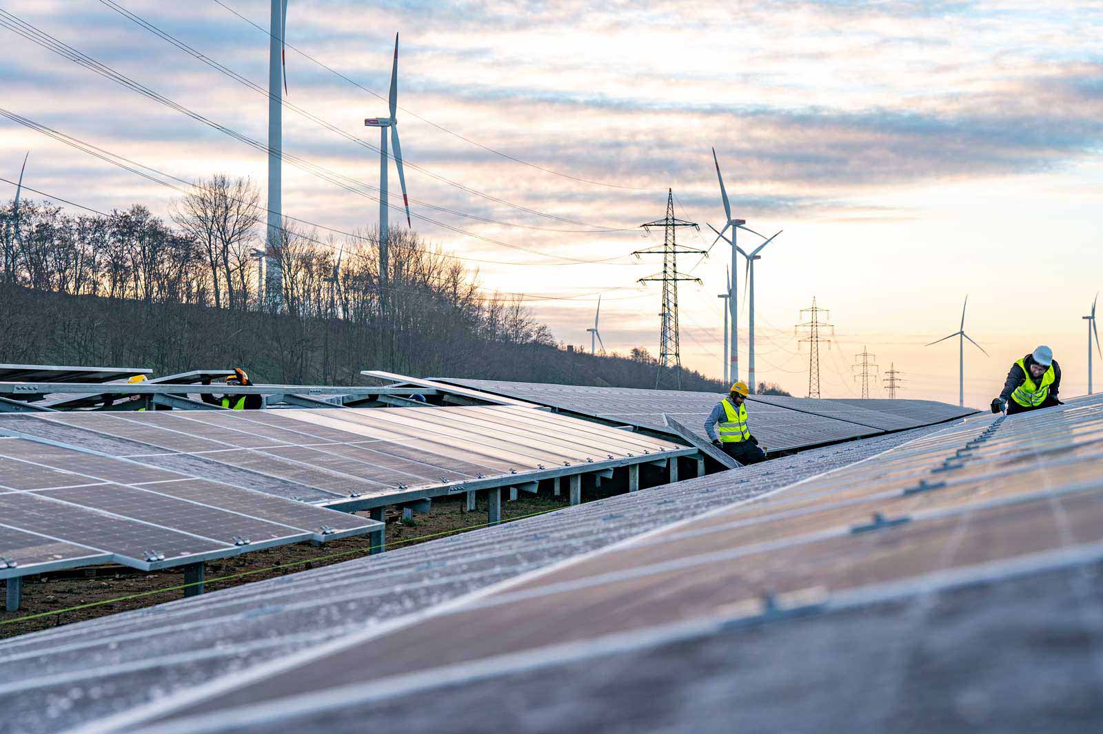 Techniker arbeiten an Solarpanels mit Windturbinen und Stromleitungen im Hintergrund während des Sonnenuntergangs.