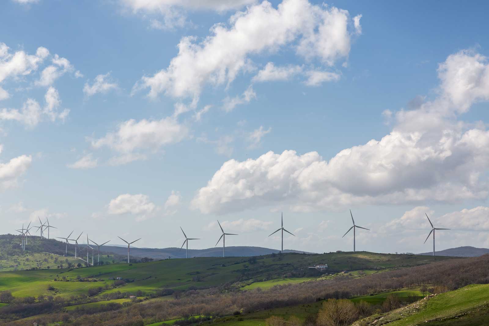 Eine malerische Landschaft mit mehreren Windkraftanlagen auf sanften grünen Hügeln unter einem blauen Himmel mit flauschigen weißen Wolken.