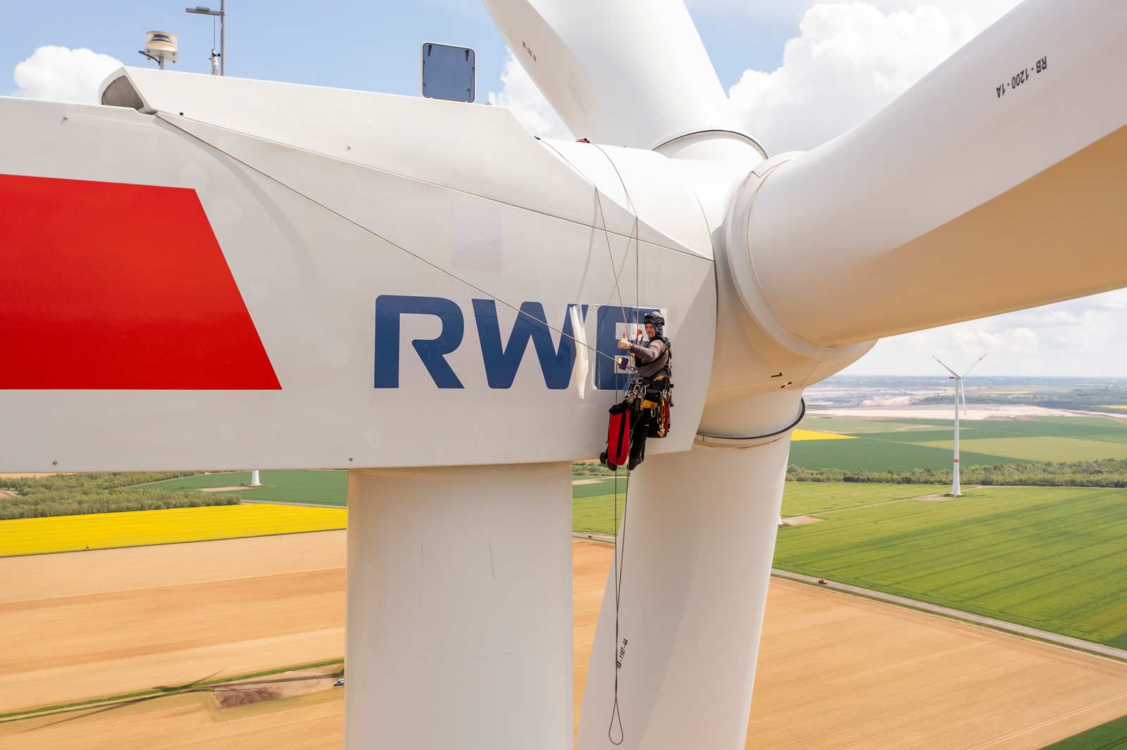 Ein Techniker seilt an einer Windkraftanlage ab, die das RWE-Logo vor einer Kulisse aus grünen und gelben Feldern zeigt.