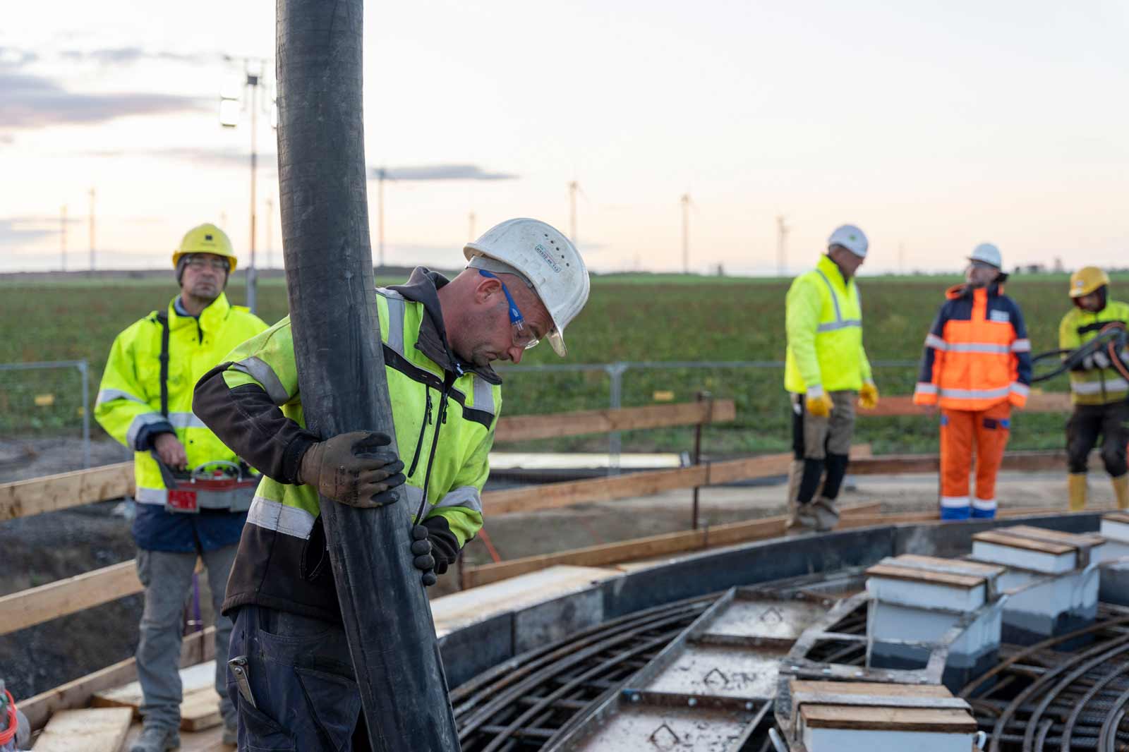 Bauarbeiter in Sicherheitskleidung gießen Beton in ein Fundament bei Sonnenuntergang, mit Windkraftanlagen im Hintergrund.
