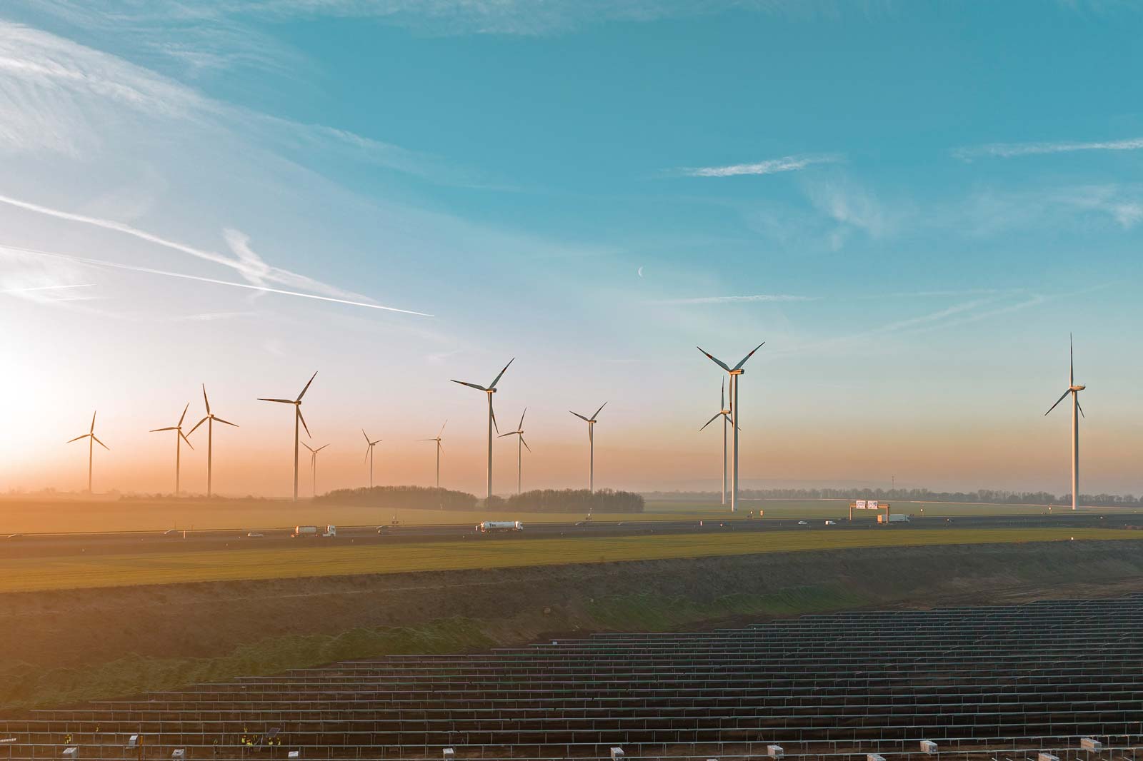 Eine Landschaft mit mehreren Windturbinen, die vor einem Sonnenaufgang und Solarpanelen im Vordergrund silhouettiert sind.
