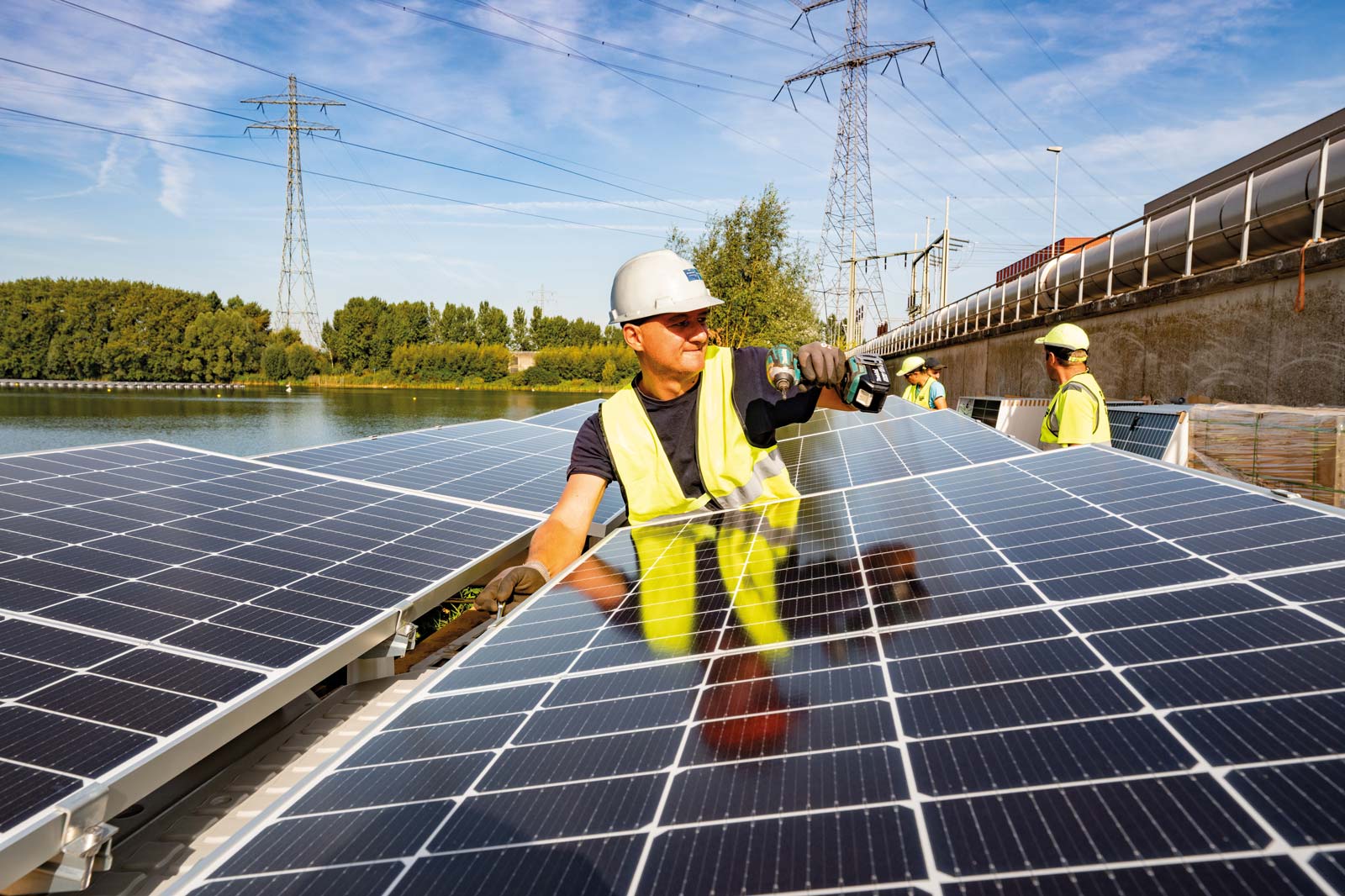 Ein Arbeiter installiert Solarpanels am Wasser, während andere Arbeiter im Hintergrund zuschauen.