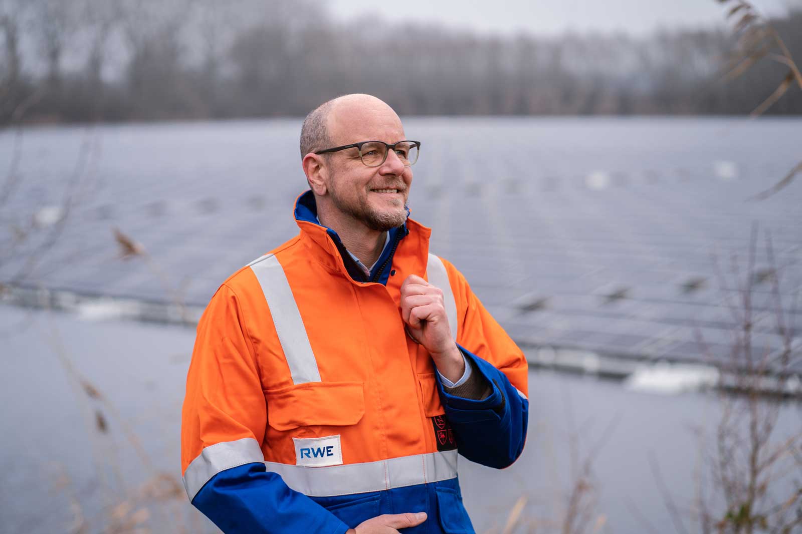 Ein Mann in einer orangefarbenen Sicherheitsjacke steht in einem wolkigen Freiluftgebiet neben Solarpanelen.