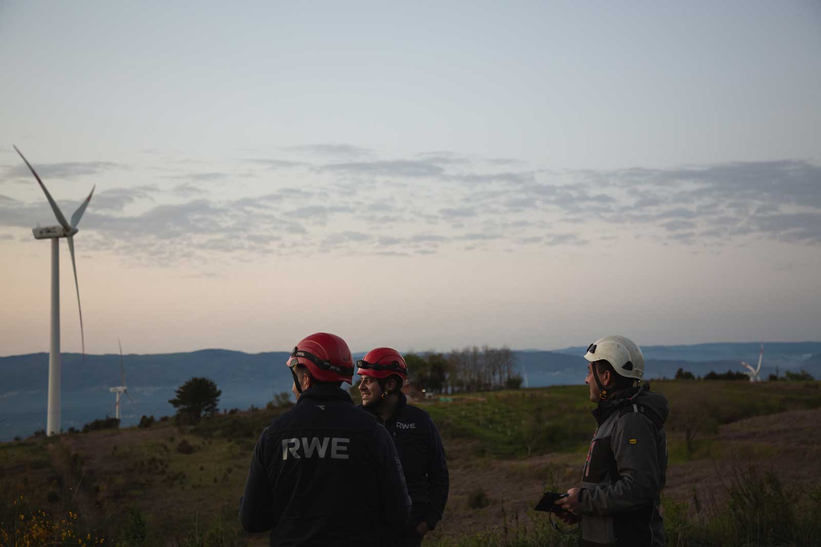 Techniker in Helmen beobachten Windturbinen vor einem Sonnenuntergang. Die Landschaft zeigt Hügel und einen bewölkten Himmel.
