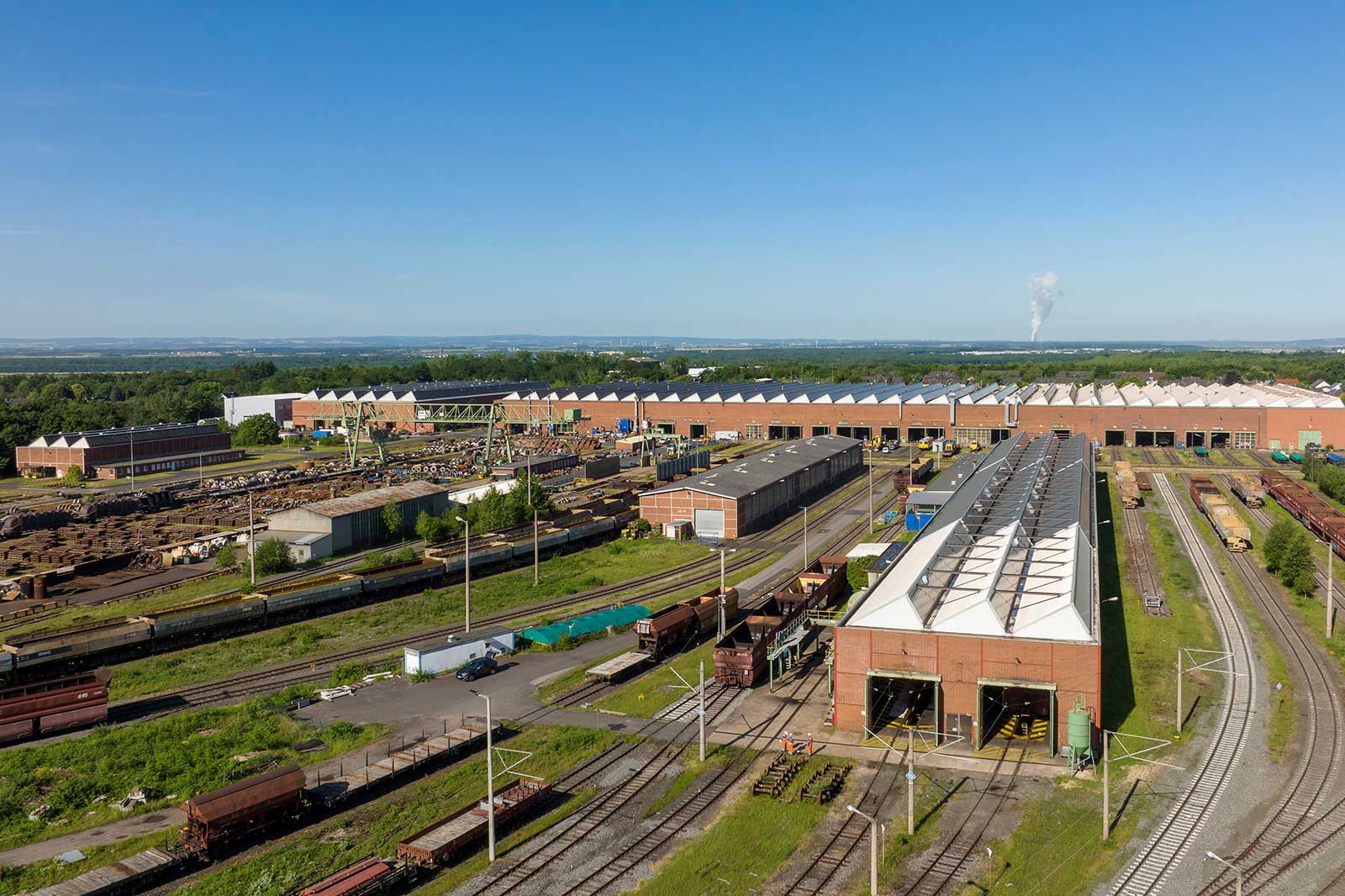 Vogelperspektive auf einen Industriehof mit Gleisen, Gebäuden und verstreuten Güterwagen unter einem klaren blauen Himmel.