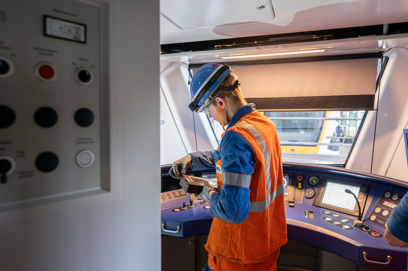 Ein Arbeiter in einer orange-blauen Uniform untersucht ein Gerät an einem Steuerpult in einer modernen Maschinenkabine.