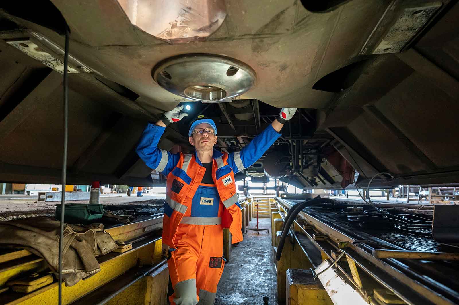 Ein Techniker inspiziert die Unterseite eines Fahrzeugs und verwendet eine Taschenlampe in einer Werkstatt voller Werkzeuge und Ausrüstung.
