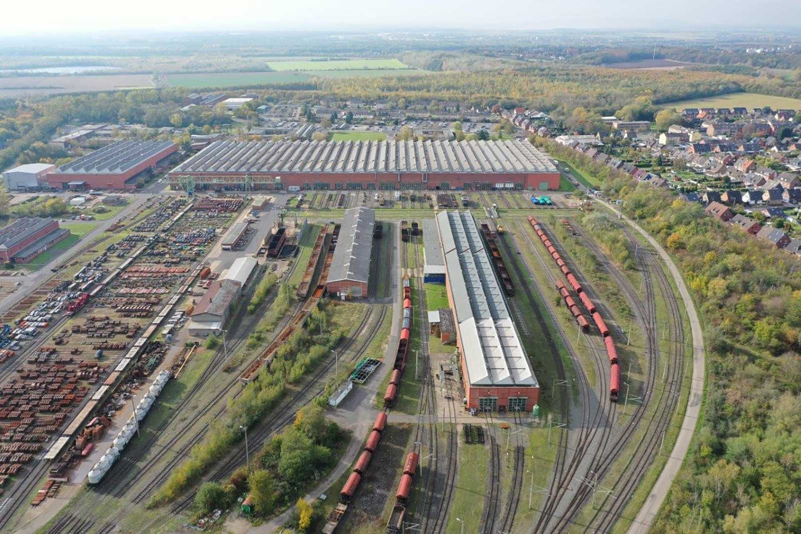 Luftaufnahme eines Bahnhofs mit mehreren Gleisen, Güterzügen und Industriegebäuden in einer ländlichen Umgebung.