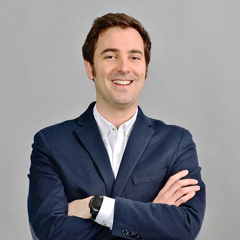 A man in a dark blazer stands with crossed arms, wearing a white shirt and a wristwatch, against a grey background.