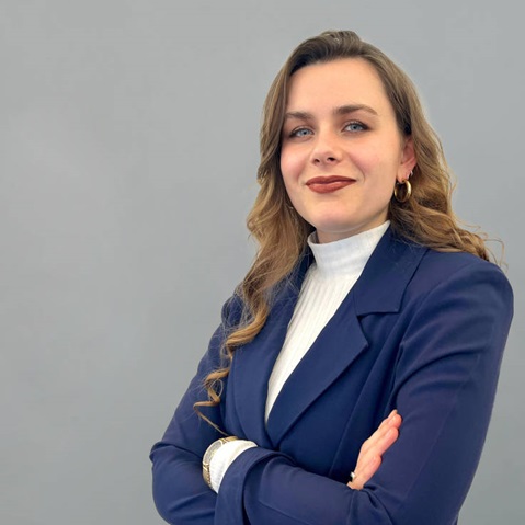 A woman in a blue blazer stands with her arms crossed in front of a grey wall.