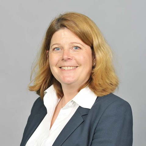 A person with blonde hair is wearing a grey jacket and a white shirt. The background is grey.