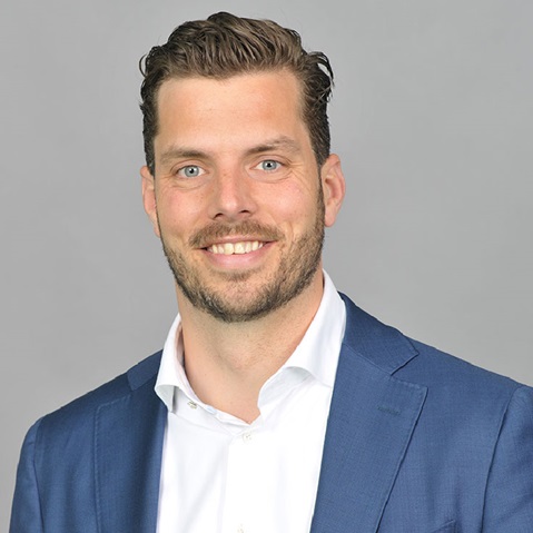 A man is wearing a blue suit and a white shirt. The background is grey.