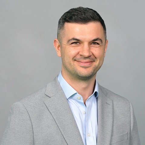 A man is wearing a grey suit and a light blue shirt. The background is grey and neutral. Portrait of Gavin Hyder.