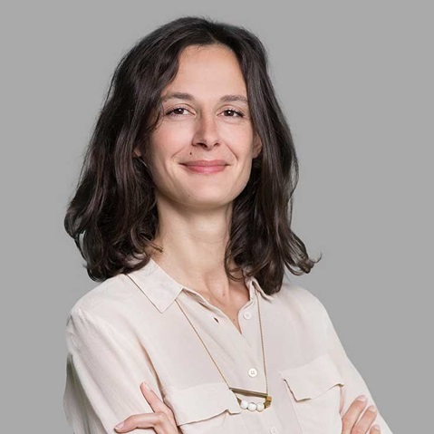 A person with shoulder-length hair wears a light shirt, smiling against a grey background.