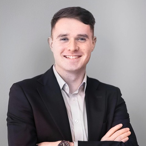 A person in a dark suit jacket and white dress shirt stands with arms crossed against a plain light gray wall. Portrait of Michael Keogh.