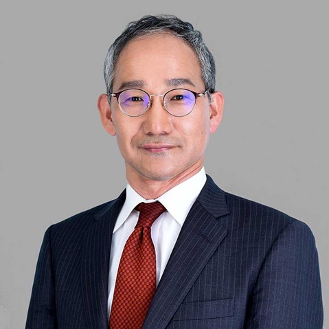 A man in a dark blue suit with a red tie, standing against a grey background.