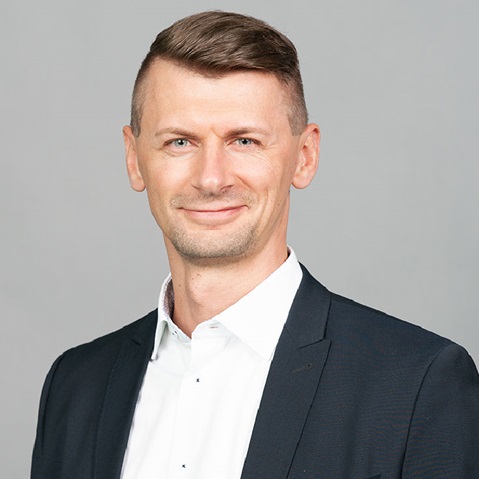 A man in a black suit and white shirt poses against a grey background, showcasing a professional appearance.