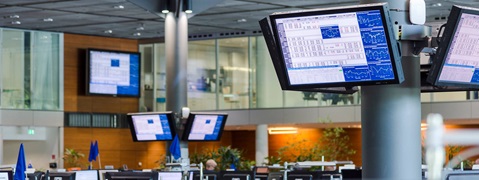 An office with multiple monitors displaying financial charts and data, featuring plants and modern architecture in the background.