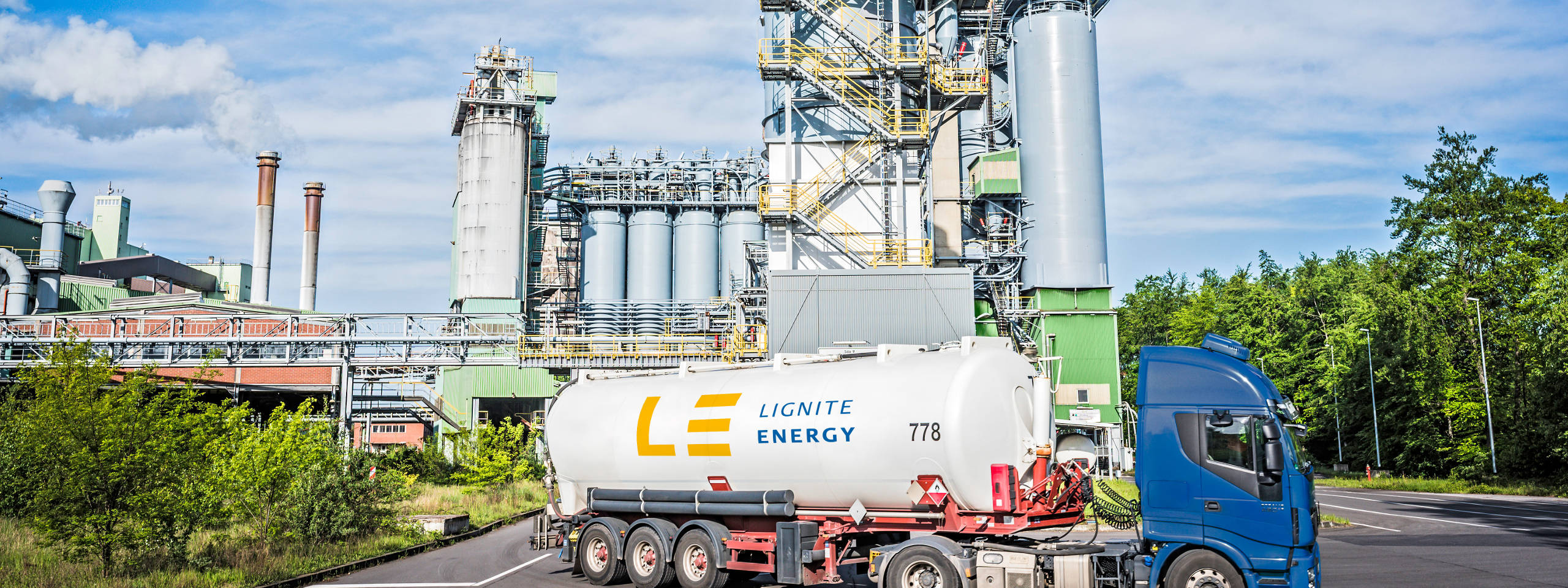 Ein Lkw mit einem Tank für Lignit Energie parkt vor einer Industriefabrik mit großen Silos und rauchenden Schornsteinen.