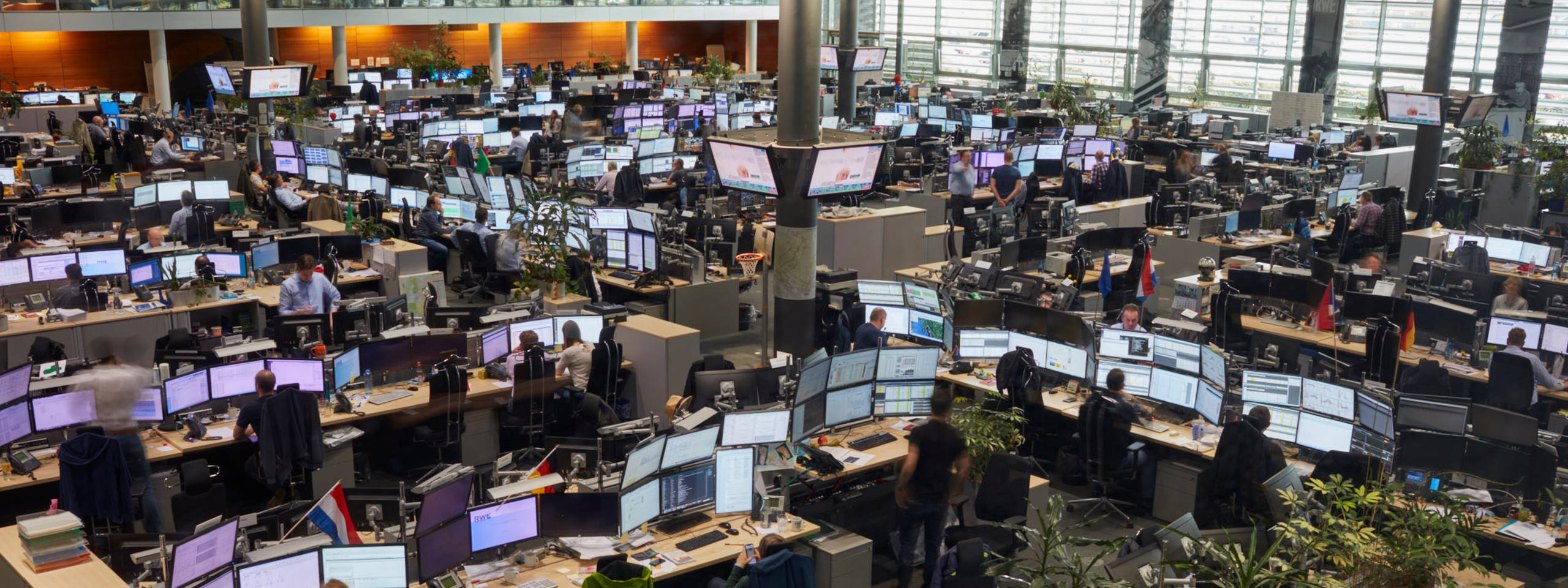 Blick auf ein modernes Büro mit zahlreichen Arbeitsplätzen und Bildschirmen, alle besetzt von Mitarbeitenden.