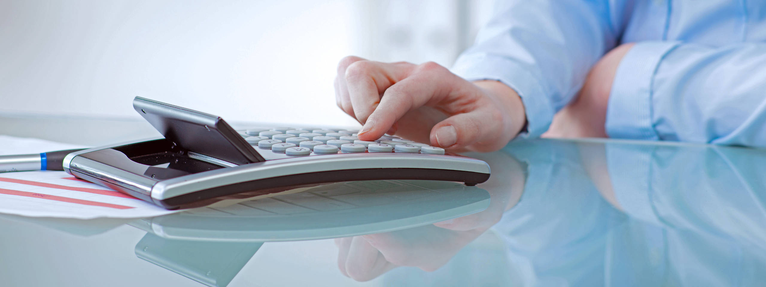 Eine Hand drückt die Tasten eines Taschenrechners auf einem Glastisch, daneben liegt ein Stift und ein Dokument.