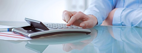 Eine Hand drückt die Tasten eines Taschenrechners auf einem Glastisch, daneben liegt ein Stift und ein Dokument.