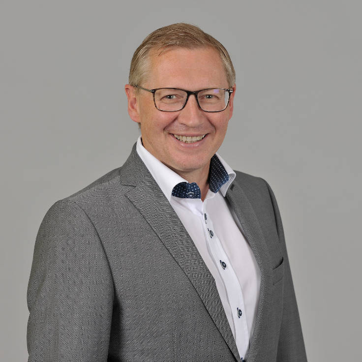 A man in a grey suit with a white shirt and blue accents stands against a grey background, portrait of Friedhelm Schillings.