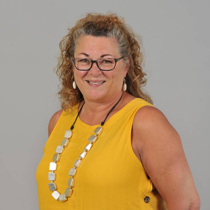 A woman with curly hair wears a yellow top and a striking necklace. The background is grey, portrait of Karen Bennett.