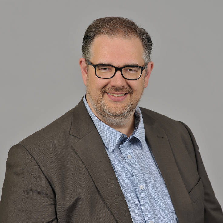 Ein Mann in einem braunen Sakko und einem hellblauen Hemd posiert vor einem grauen Hintergrund, Portrait von Ralf Rinsch.