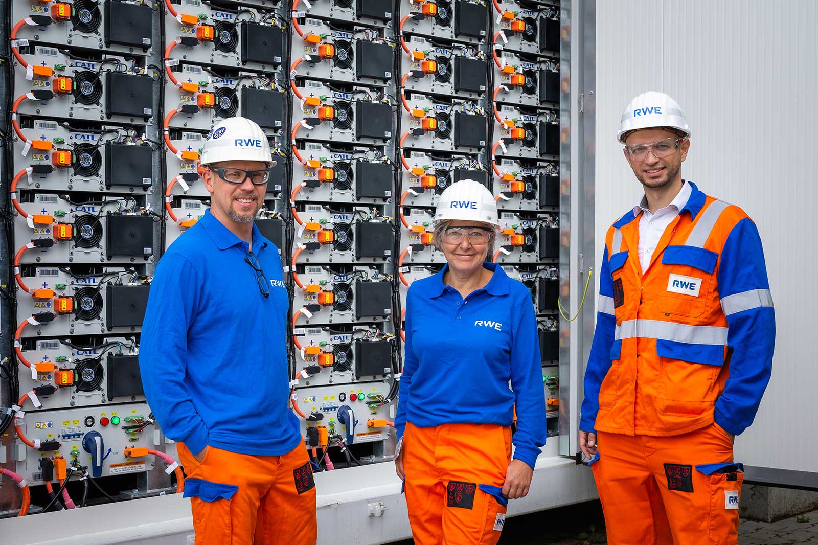 Drei Arbeiter in blauen und orangefarbenen Overalls stehen vor einer Wand mit Batteriemodulen und elektrischen Komponenten.
