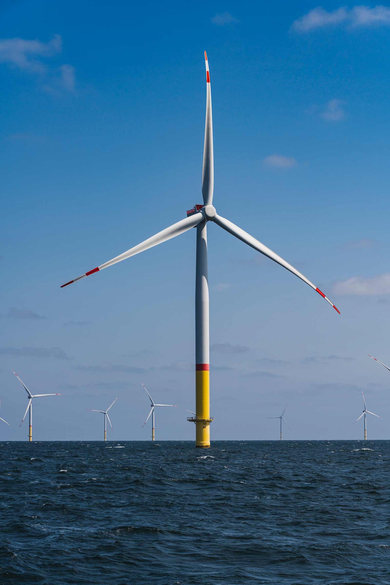 A large wind turbine stands in the water, with a blue sky and several additional turbines in the background.