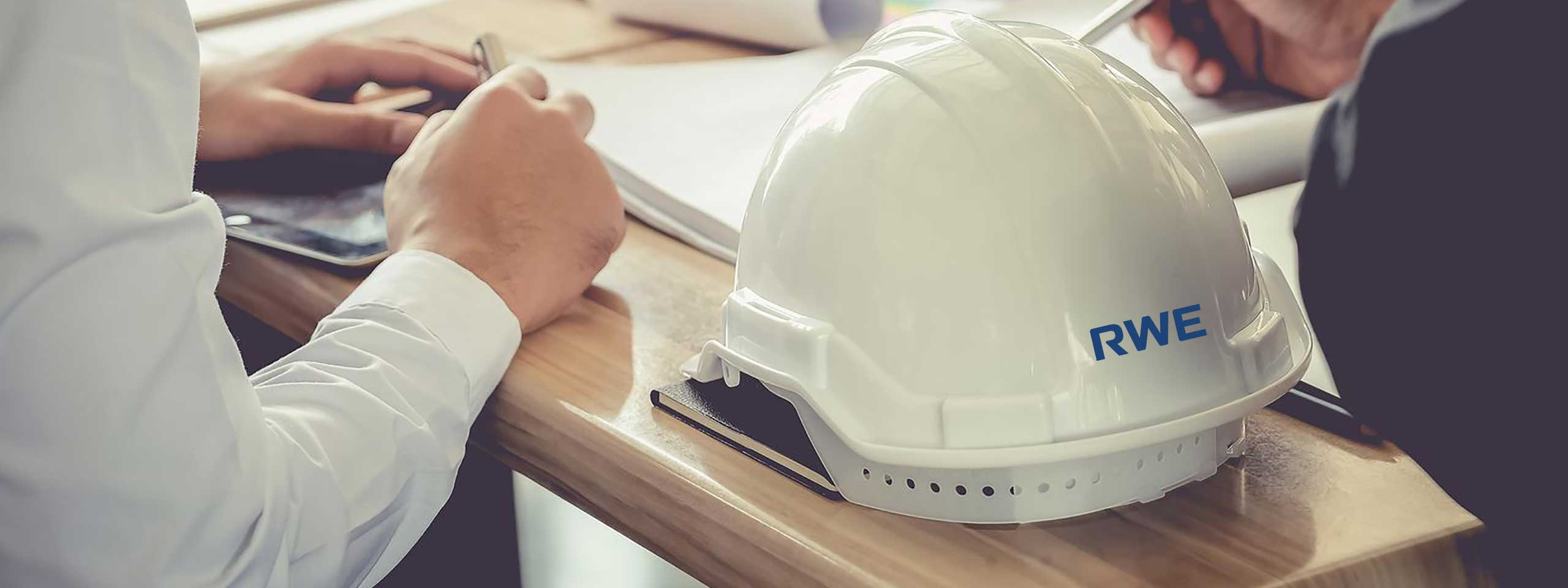 A white hard hat with 'RWE' logo on a wooden table, with hands writing and a tablet in the background.