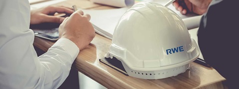 Ein weißer Helm mit dem Logo 'RWE' auf einem Holztisch, mit Händen, die schreiben, und einem Tablet im Hintergrund.