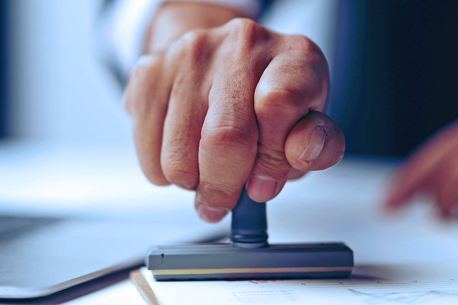 A hand pressing a stamp onto a sheet of paper. The background is blurred, creating an office atmosphere.