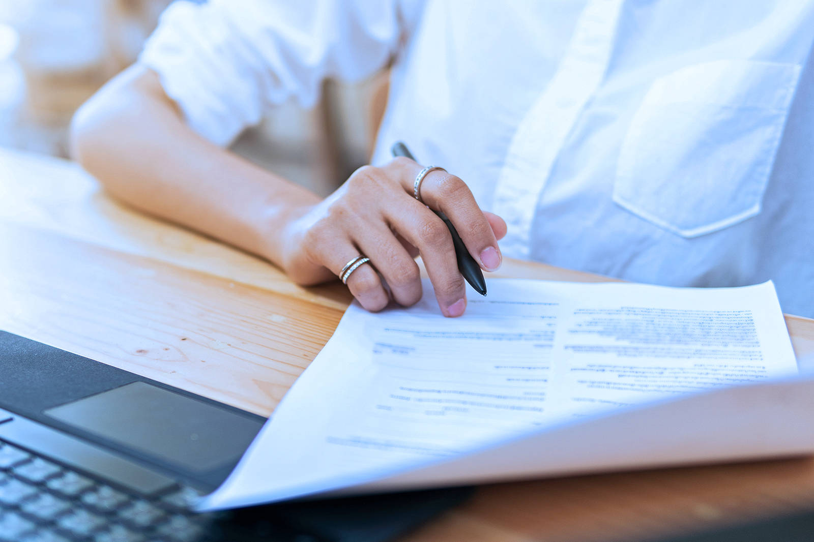 A person holds a pen and examines a document on a wooden table next to a laptop.