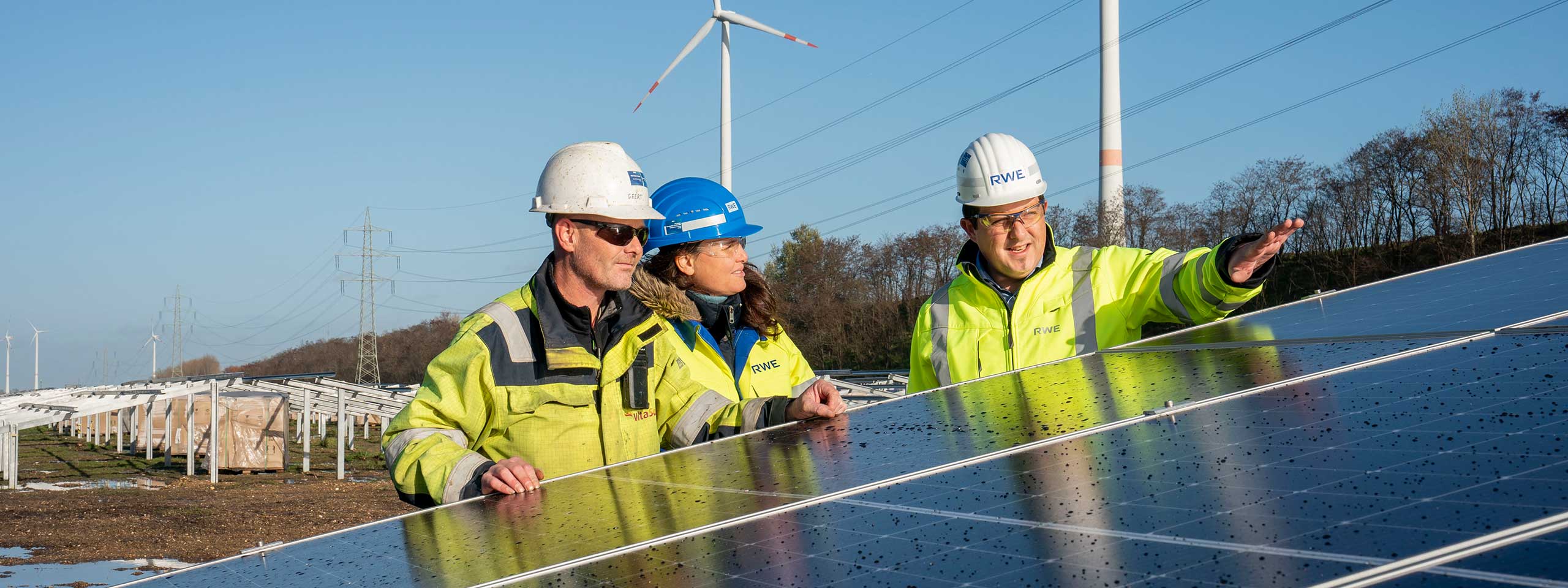Three professionals in safety gear are standing next to solar panels in a field with wind turbines in the background.