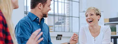 Three people are conversing in a modern office. A blonde woman and a man in a blue shirt are visible.