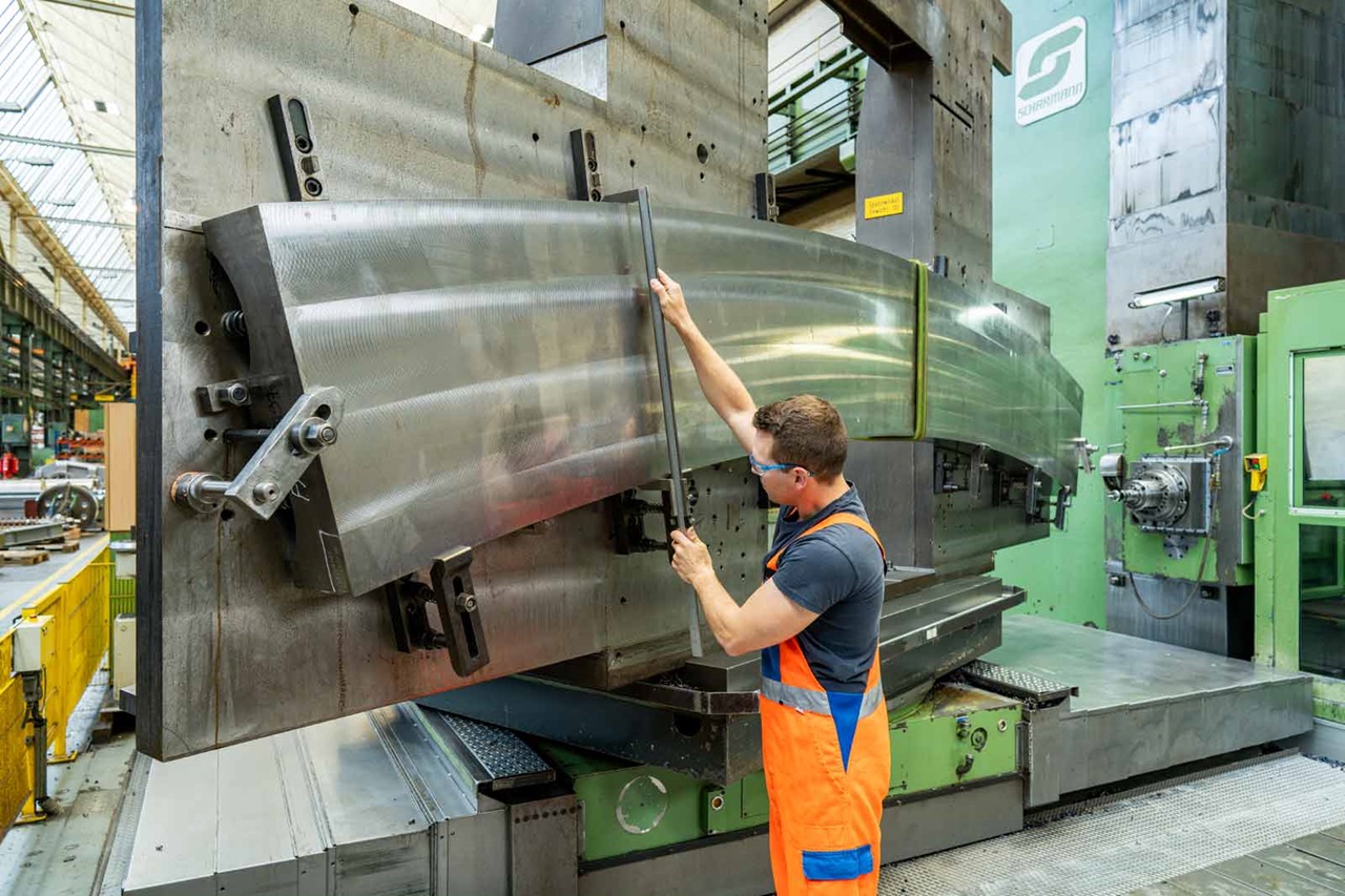 A worker measures a large metal component in a factory; precise machinery is visible in the background.