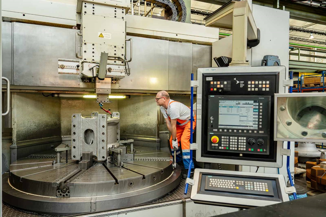 A worker operates a large lathe machine in a factory while processing a metal component.