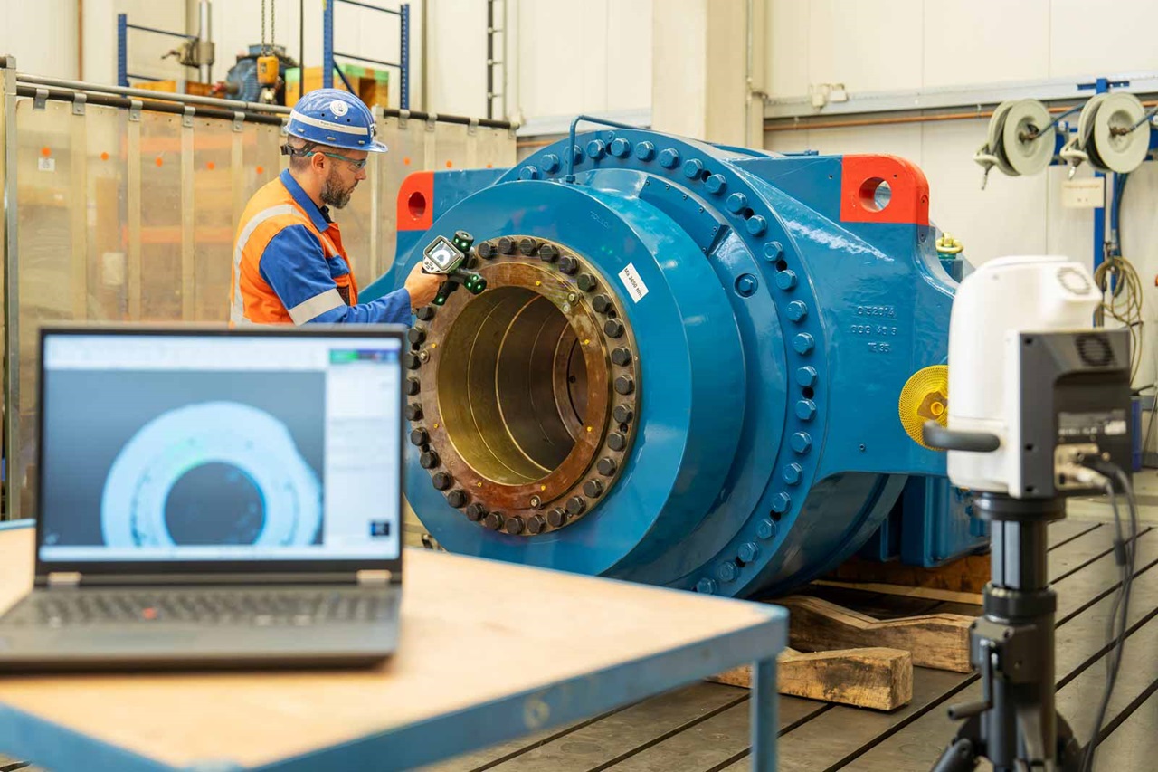 A worker inspects a large blue machine using a measuring device. A laptop displays a technical drawing of the machine.